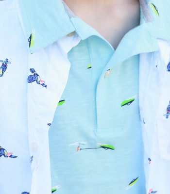 a child wearing a blueish green polo shirt with boats on it and a white button up shirt with toucans layered over it