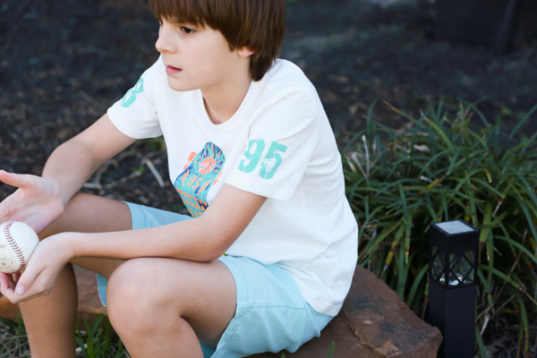 a little boy sitting down holding a baseball wearing a white tshirt and blueish colored shorts - perfect spring styles at oshkosh b'gosh